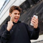 Cheerful man holding smartphone and celebrate win a bet. Soccer bet, sports gambling. Football stadium on the background.